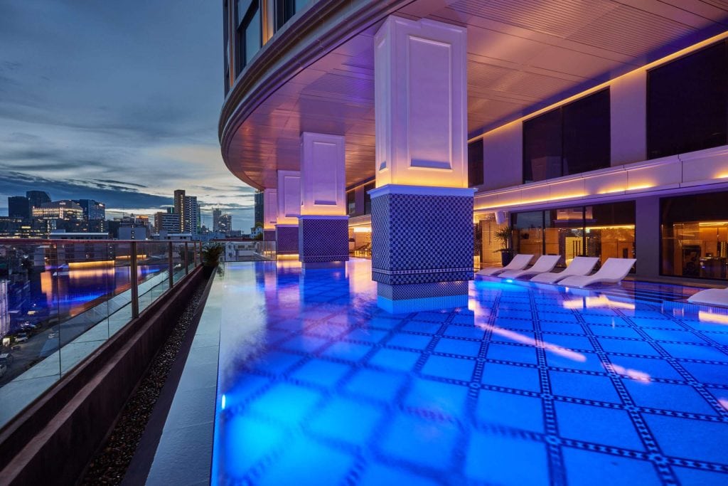 A modern rooftop pool glows with blue lights at dusk, surrounded by lounge chairs and columns. City buildings and a cloudy sky are visible in the background through glass railings.