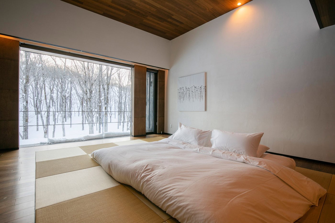 Minimalist bedroom with a low bed on tatami mats, large window overlooking a snowy forest, white walls, wooden ceiling, and simple wall art, creating a serene and peaceful atmosphere.