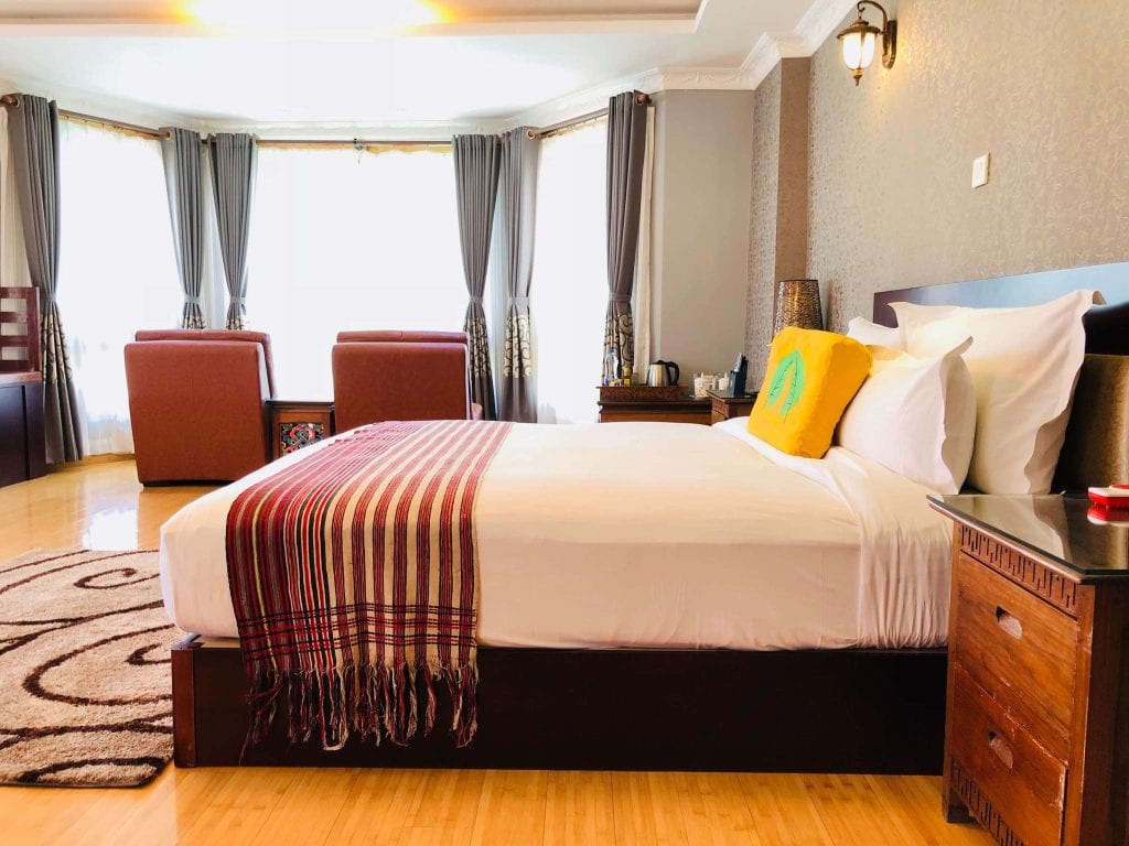 A spacious, well-lit bedroom with a large bed covered in white linens, a striped throw, and a yellow pillow. There are brown armchairs by the window, gray curtains, wooden furniture, and a patterned rug on the floor.