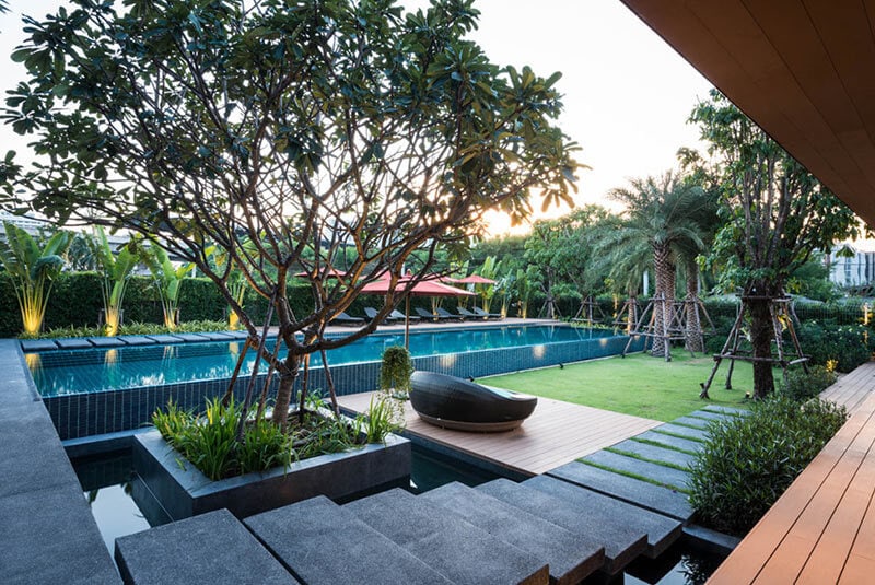 A modern backyard with a large swimming pool surrounded by greenery, palm trees, stepping stones, a wooden deck with a lounge chair, and landscaped garden in the evening light.