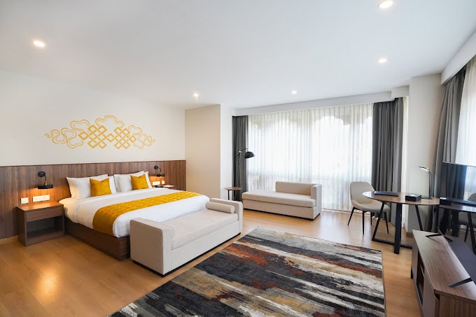 A spacious hotel room features a large bed with yellow accents, a white sofa, a desk with a chair, floor lamps, and a modern patterned rug. Large windows with sheer curtains let in natural light.