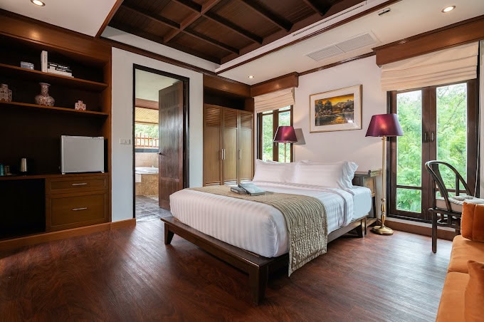 A modern hotel room with a neatly made double bed, wooden floors, large windows with beige shades, two purple lamps, a bookshelf, and a door leading to a bathroom. The room is bright and surrounded by greenery outside.