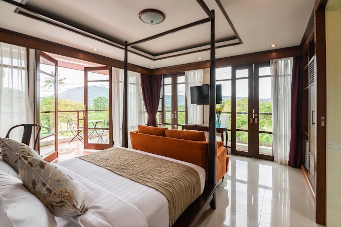 A spacious, modern bedroom with a four-poster bed, orange sofa, large windows, and glass doors opening to a balcony with mountain and greenery views. Natural light fills the room, creating an airy atmosphere.