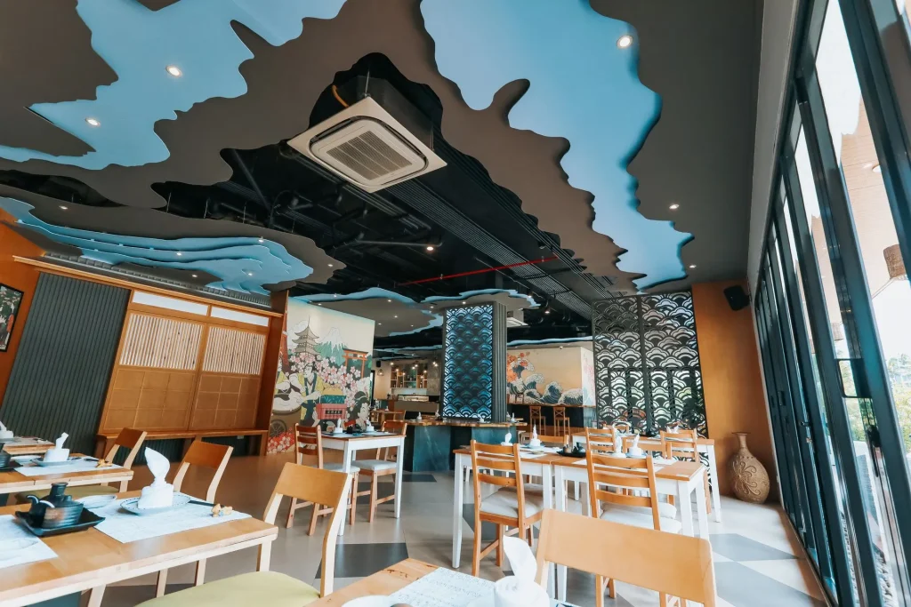 A modern restaurant interior with wooden tables and chairs, unique blue and black cloud-like ceiling design, large windows, and artistic wall murals in the background. Place settings are neatly arranged on the tables.