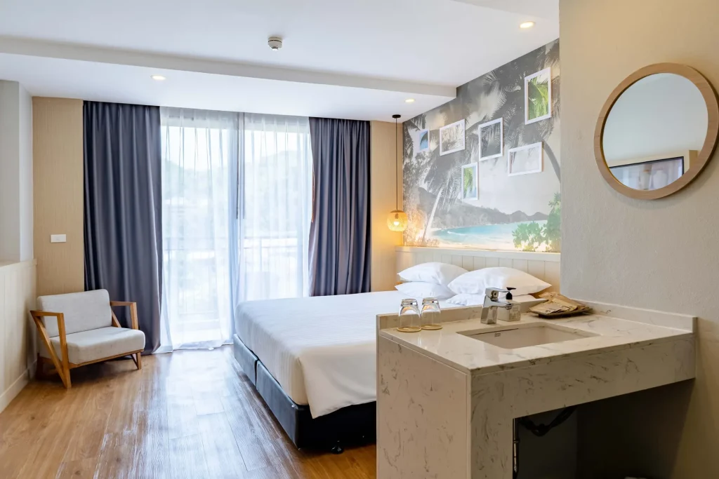 A modern hotel room with a large bed, light wood floors, a small armchair, a sink with toiletries, and a round mirror. Large windows with sheer curtains let in natural light. A decorative wall features photos of nature.