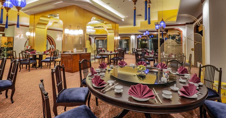 Elegant restaurant interior with round tables set with purple napkins and white dishes, dark cushioned chairs, warm lighting, blue hanging lamps, and mirrored golden walls creating a sophisticated atmosphere.