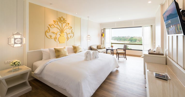 Bright, modern hotel room with a large bed, white bedding, gold accents, a sitting area by a window overlooking greenery and water, a TV on the wall, and stylish light fixtures.