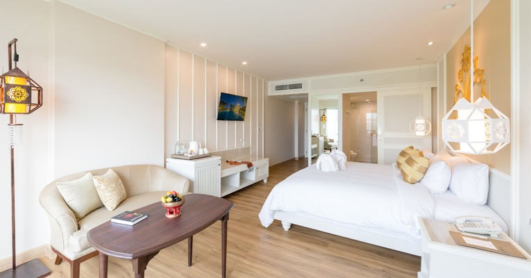Bright, modern hotel room with a large bed, white bedding, a small round table with a fruit bowl, a beige sofa, and wall decor. The room has wooden flooring and an open area leading to the bathroom.