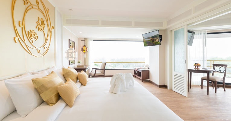 A bright, spacious hotel room with a large bed, gold and white decor, folded towel swans on the bed, a sitting area by wide windows, and a dining table near a balcony with natural light streaming in.