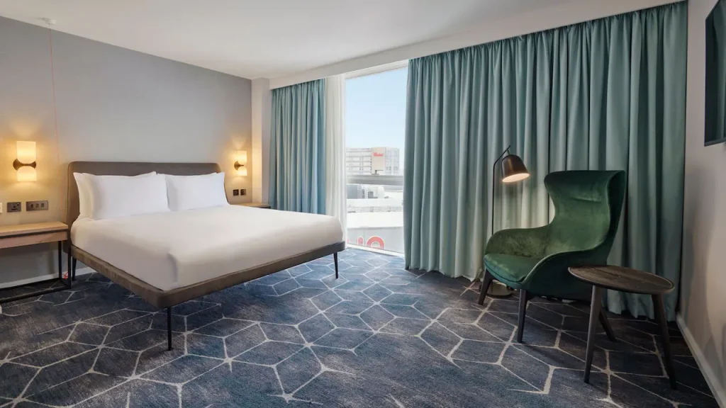 Modern hotel room with a bed, white linens, bedside tables, wall lamps, a green armchair with a floor lamp, round side table, blue patterned carpet, and large window with teal curtains showing city buildings outside.