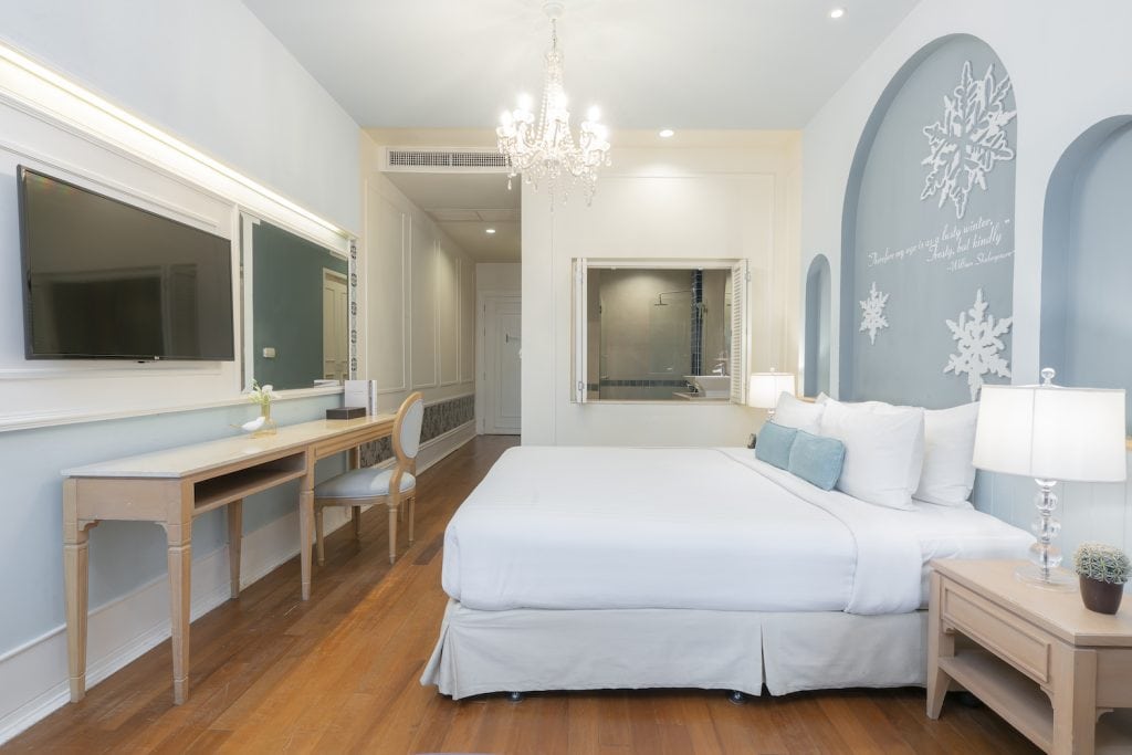 A bright, modern hotel room with a large bed, white bedding, blue accent pillows, a wooden desk and chair, a flat-screen TV, chandelier, bedside table with lamp, and snowflake decorations on a blue accent wall.