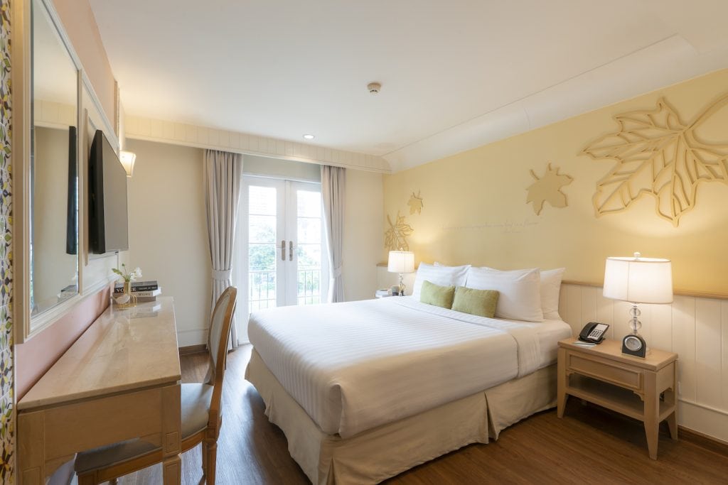A bright hotel room with a large bed, two bedside tables with lamps, a wall-mounted TV, a desk with a mirror, and double doors leading to a balcony. The decor is minimalist with light-colored walls and wooden floors.