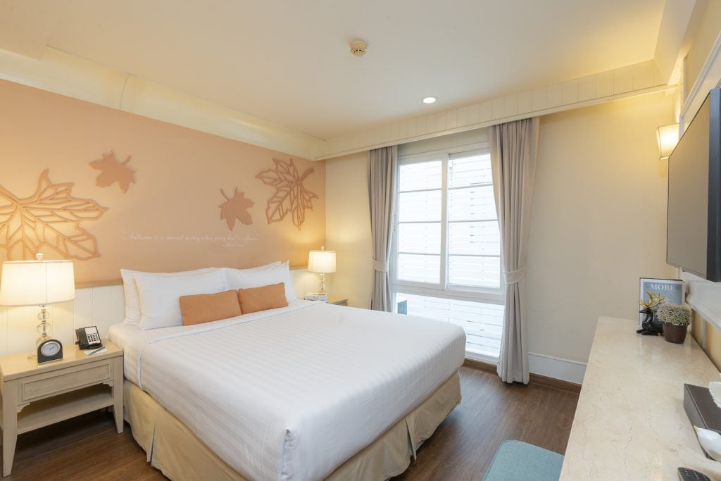 A bright hotel room with a large bed, two bedside tables with lamps, wall art with leaves and turtles, a window with curtains, a flat-screen TV, and a desk with a chair.