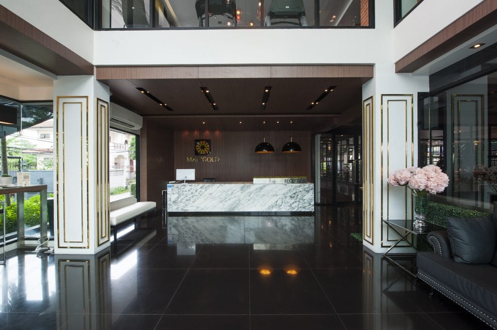Modern hotel lobby with a marble reception desk, black shiny floor, cushioned benches, large windows, and decorative white and gold columns. Pink flowers are arranged on a side table, and three pendant lights hang above the desk.
