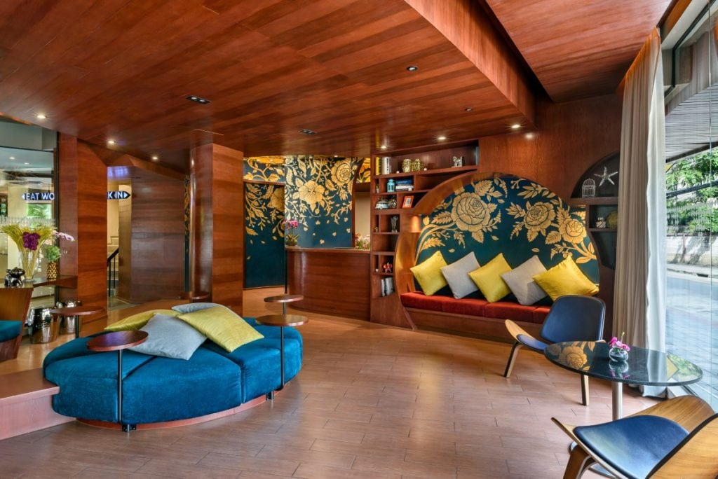 A modern hotel lobby with wooden walls and ceiling, a blue and gold floral mural, a built-in red cushioned bench with colorful pillows, a blue circular sofa, glass tables, and large windows letting in natural light.