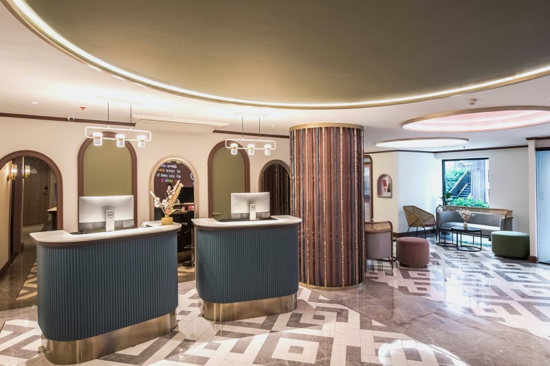 Modern hotel lobby with two reception desks, contemporary lighting, geometric tile flooring, a round column, and a seating area with chairs and a window in the background. The space has a stylish and welcoming design.