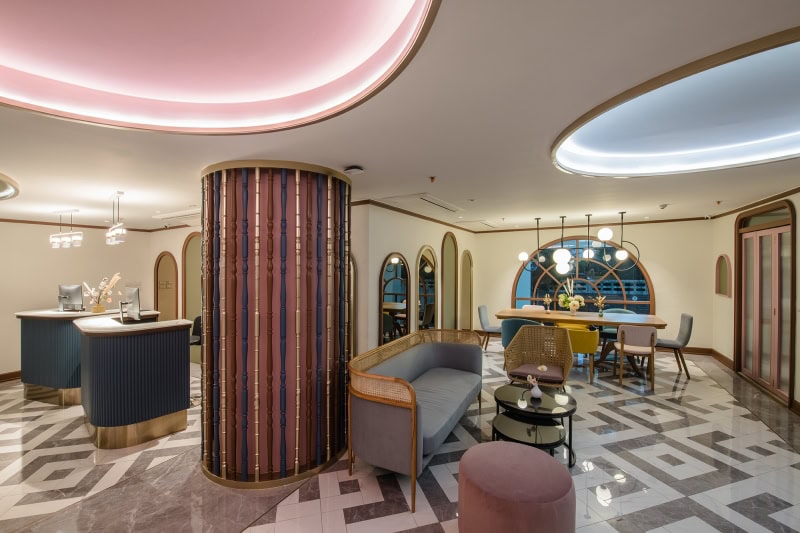 A modern, stylish lounge with geometric patterned floor, contemporary seating, curved ceiling lights, and large arched windows. The space features a mix of soft pastel colors and metallic accents.