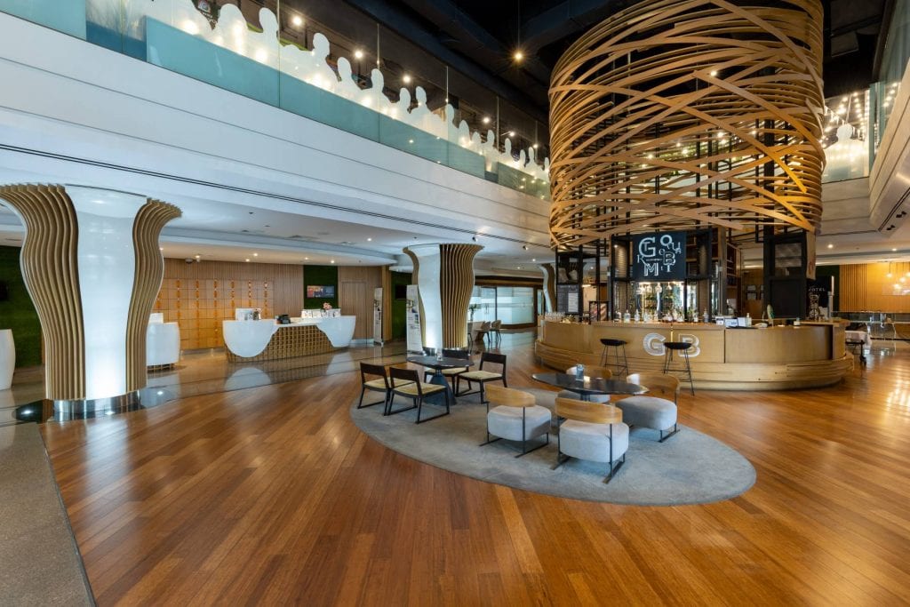 A spacious, modern hotel lobby with wood floors, unique spiral wooden structures, a stylish café counter, lounge seating, and natural light from floor-to-ceiling windows.