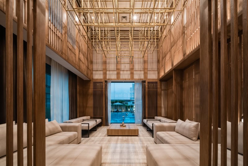 A modern lounge with beige sofas and a wooden table, enclosed by vertical wooden slats and a geometric, gold ceiling feature. Large windows at the end show an outdoor pool area. The space has warm lighting and a spacious feel.