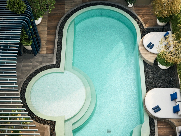 Aerial view of a modern swimming pool with a small circular shallow area attached, surrounded by lounge chairs with blue cushions and greenery on a deck.
