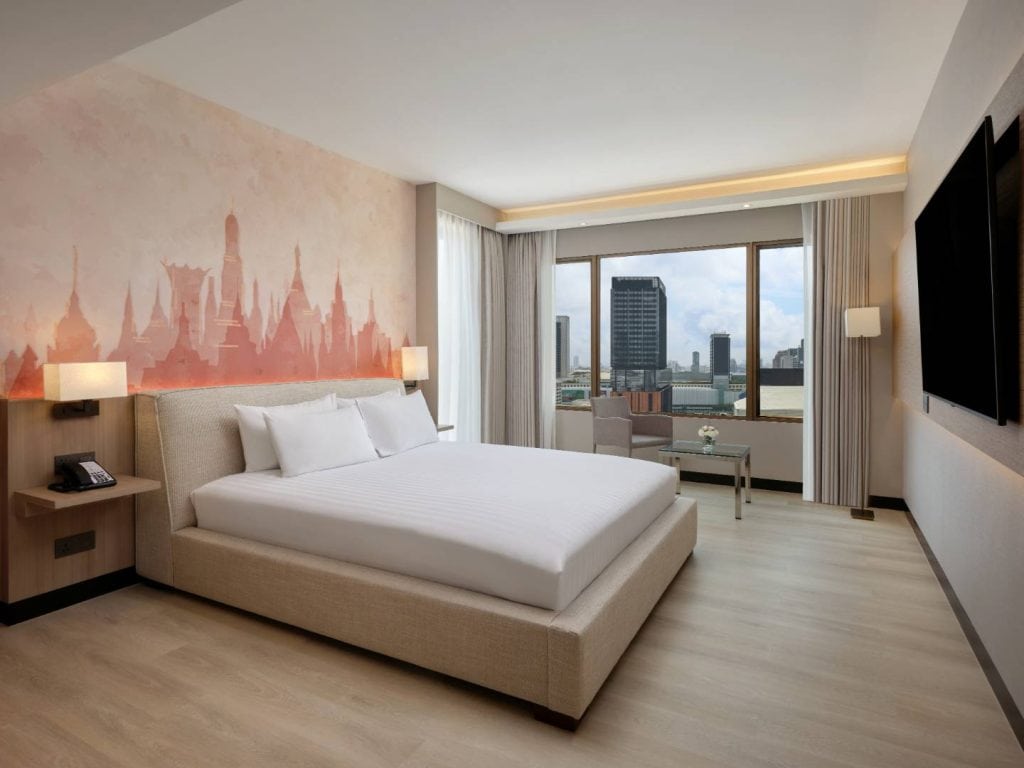 Modern hotel room with a large beige bed, light wood floors, wall art behind the bed, two armchairs by a window overlooking a river and city buildings, and a mounted flat-screen TV on the opposite wall.