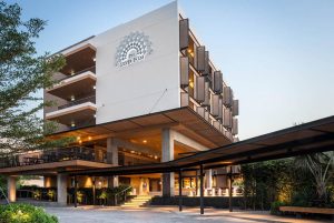 A modern hotel building with a geometric facade and open balconies, featuring the Silver Palm logo on the front. The entrance is surrounded by greenery and well-lit in the early evening.