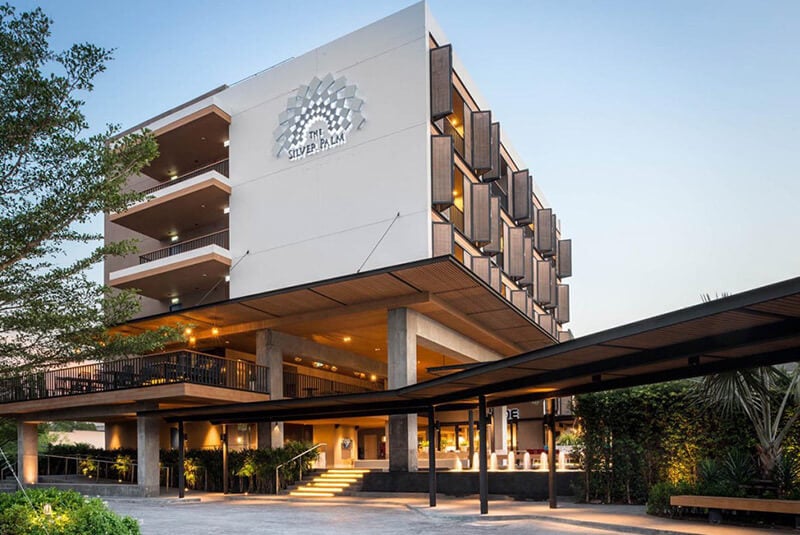 A modern hotel building with a geometric facade and open balconies, featuring the Silver Palm logo on the front. The entrance is surrounded by greenery and well-lit in the early evening.