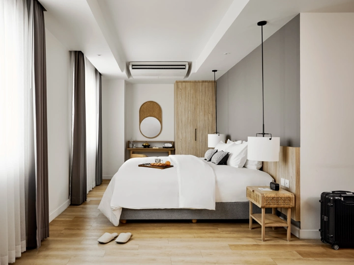 A modern, minimalistic hotel room with a large bed, white bedding, pendant lights, wooden furniture, a woven stool, a suitcase, and slippers on a wooden floor. Soft natural light enters through tall windows with curtains.