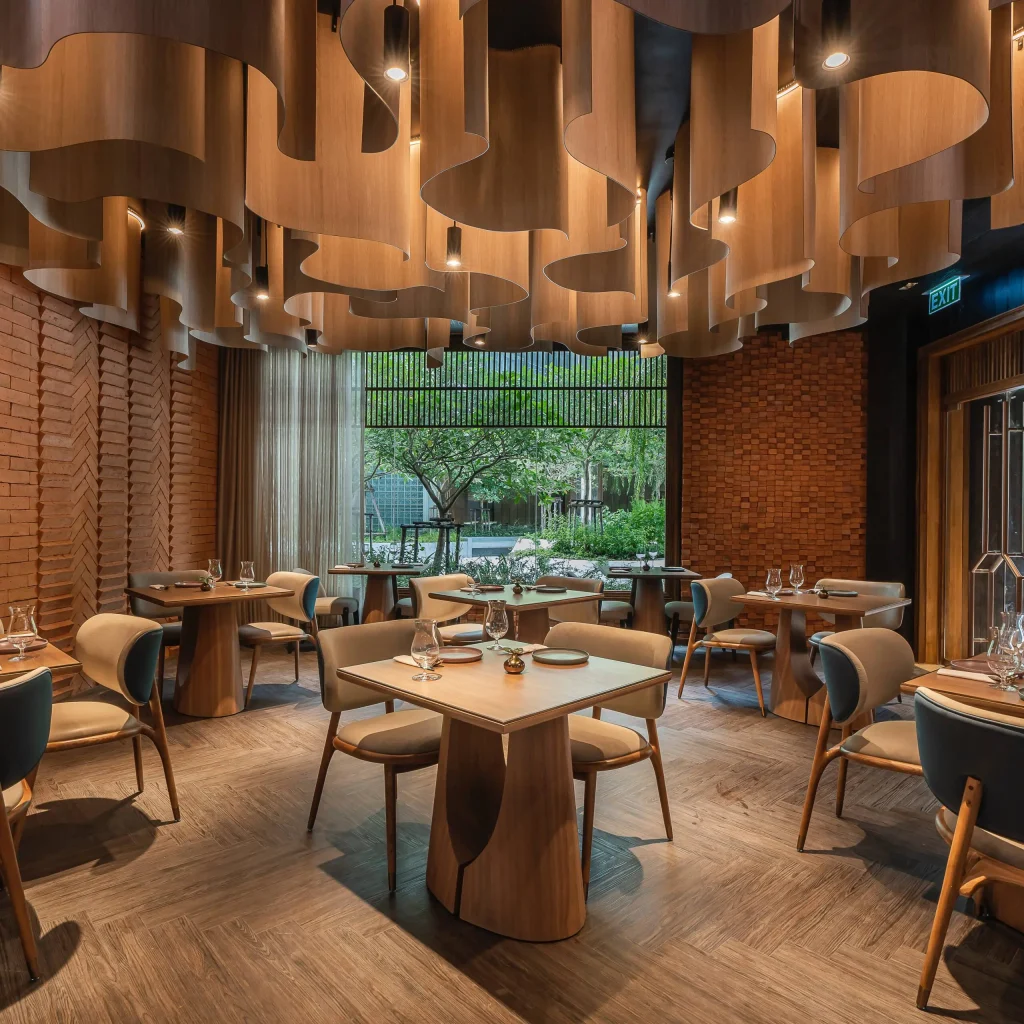 A modern restaurant interior with wooden tables and chairs, unique sculptural wooden ceiling panels, brick walls, and large windows offering a view of greenery outside. The space is elegantly set for dining.