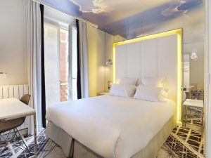 A modern hotel room with a double bed, white bedding, illuminated headboard, mirrored wall, desk and chair, large window with curtains, and a ceiling mural of clouds and blue sky.