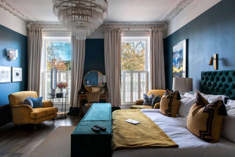Elegant bedroom with teal walls, large bed with mustard and teal pillows, mustard armchair, chandelier, two tall windows with white shutters, long curtains, and wall art. Natural light fills the cozy, stylish space.