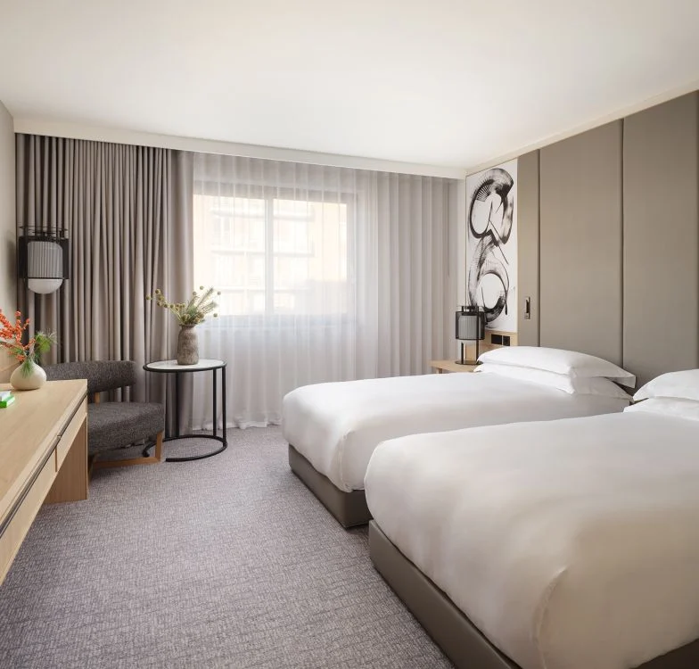 Modern hotel room with two double beds, neutral tones, a window with sheer curtains, a wooden desk, a chair with a small round table holding a vase, and minimalistic wall art above the nightstand.