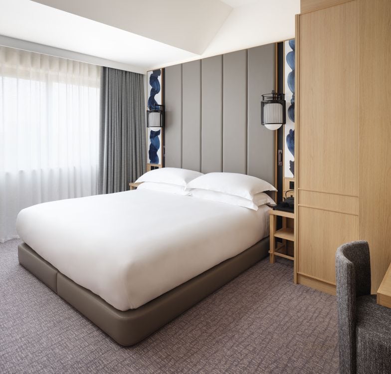 A modern hotel room with a neatly made double bed, light gray walls, a wooden wardrobe, bedside tables, and large window with sheer curtains letting in natural light.