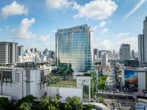 A tall glass Novotel hotel building stands amid busy city streets and high-rise buildings under a partly cloudy sky in an urban setting.