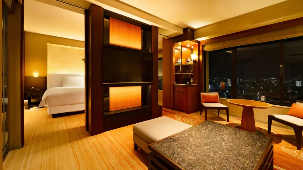 Elegant hotel suite with warm lighting, featuring a bedroom with a white bed, a sitting area with chairs and a table, a marble-topped coffee table, wooden accents, and large windows overlooking a city skyline at night.