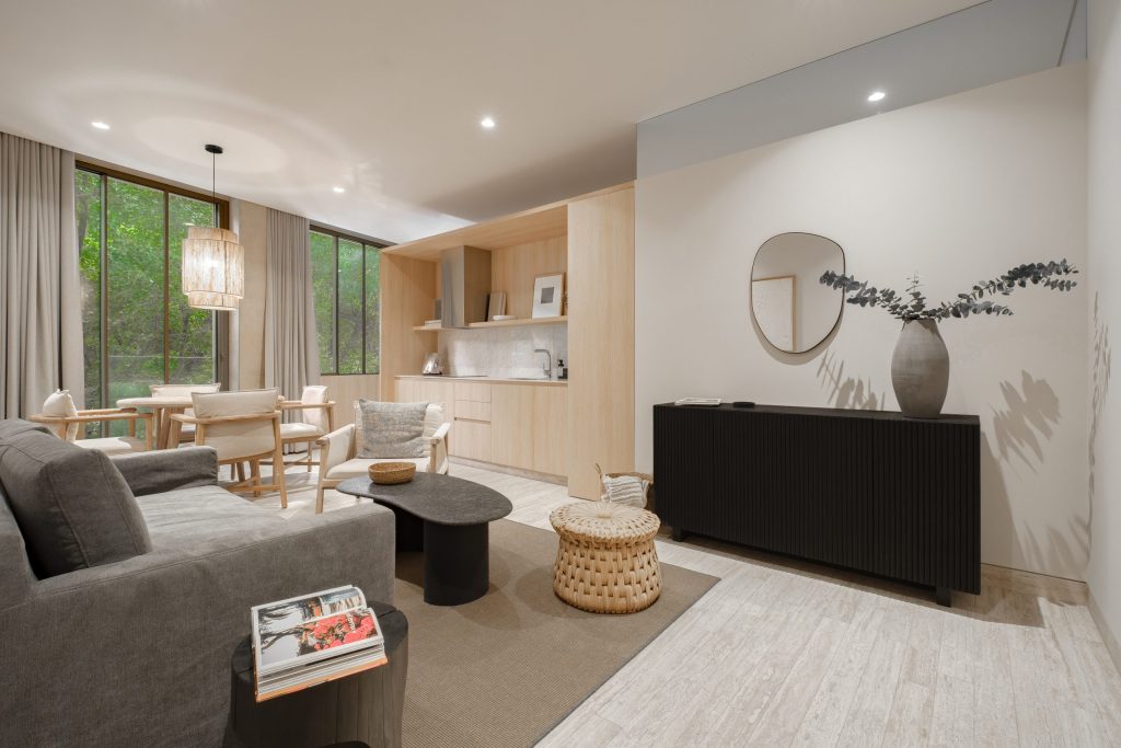 Modern living room and kitchen with neutral tones, gray sofa, black coffee table, wicker stool, dining table by large window with greenery outside, and a sideboard with a round mirror and decorative vase.
