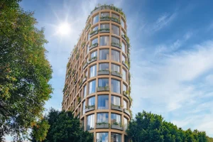 A modern, multi-story building with large windows and balconies lined with green plants, set against a blue sky with scattered clouds and the sun shining, partially framed by trees in the foreground.