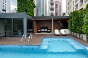 A modern outdoor pool with a wooden deck, lounge chairs, and greenery, surrounded by tall city buildings and contemporary architectural features.