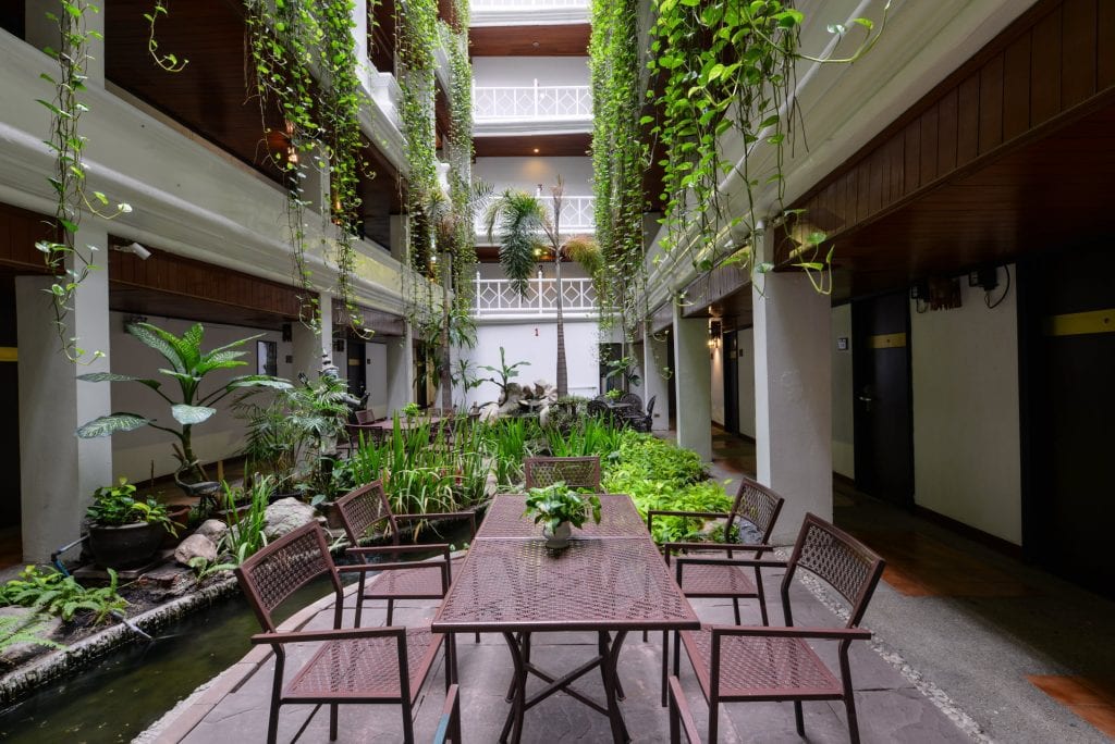 A lush indoor courtyard with hanging vines, a central garden, a rectangular table with six chairs, and surrounding balconies, creating a tranquil and inviting atmosphere.