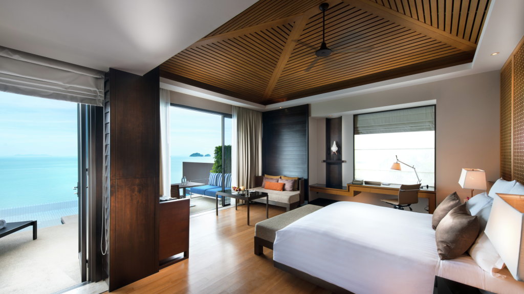A modern hotel room with a large bed, seating area, desk, and floor-to-ceiling windows offering an ocean view; a terrace with an infinity pool is visible outside.