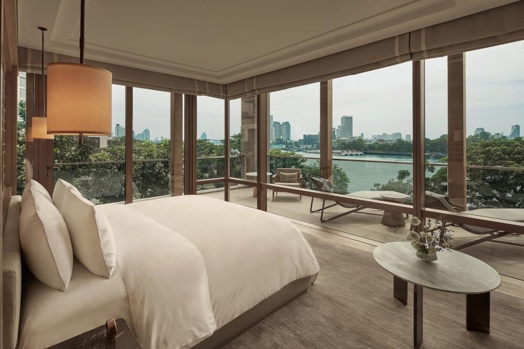 A modern bedroom with large windows offering a city skyline and river view, featuring a double bed, lounge chairs on a balcony, and a small table with a flower vase.