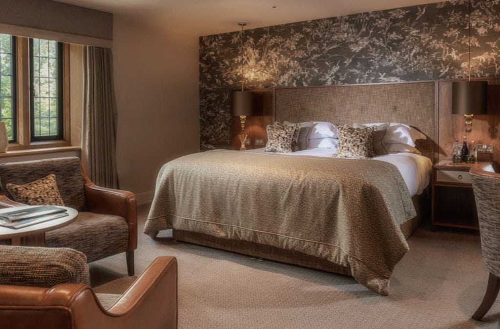A cozy, elegant hotel room with a large bed, patterned headboard, two bedside tables with lamps, a sitting area with armchairs and a small table, neutral-toned decor, carpeted floor, and a window with drapes.