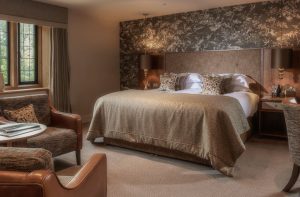 A cozy, elegant hotel room with a large bed, patterned headboard, two bedside tables with lamps, a sitting area with armchairs and a small table, neutral-toned decor, carpeted floor, and a window with drapes.