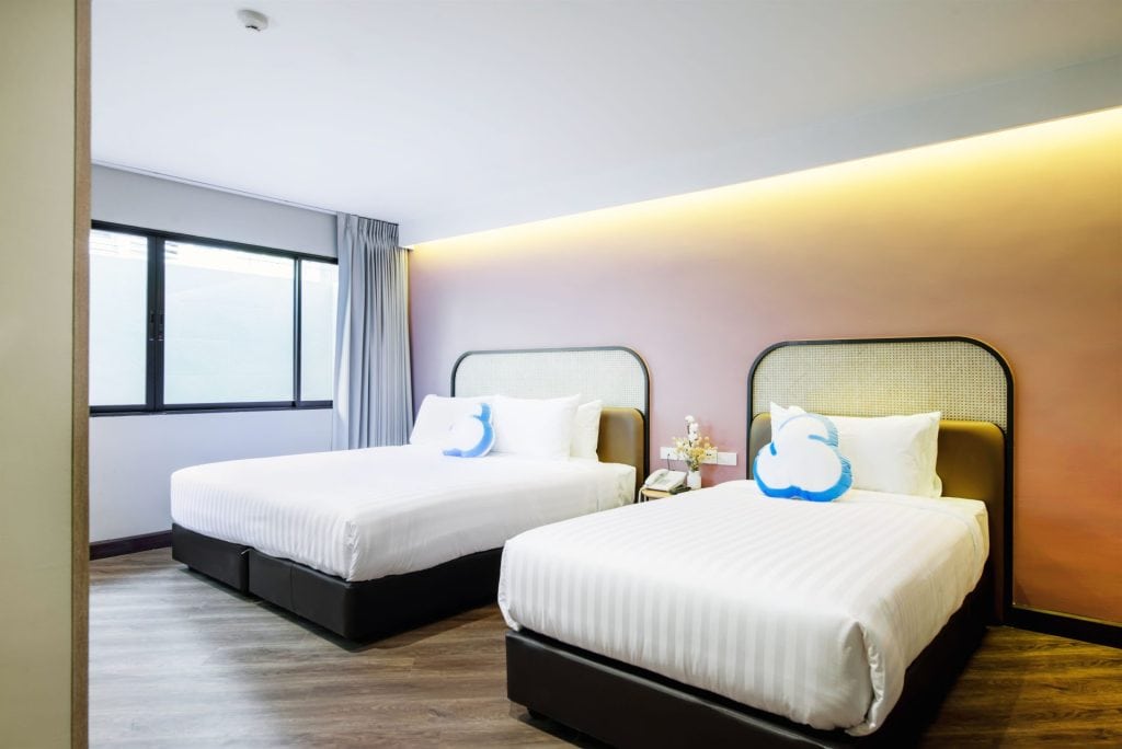 Modern hotel room with two double beds, white bedding, cloud-shaped pillows, a bedside table with flowers, wood flooring, and a large window with gray curtains letting in natural light.