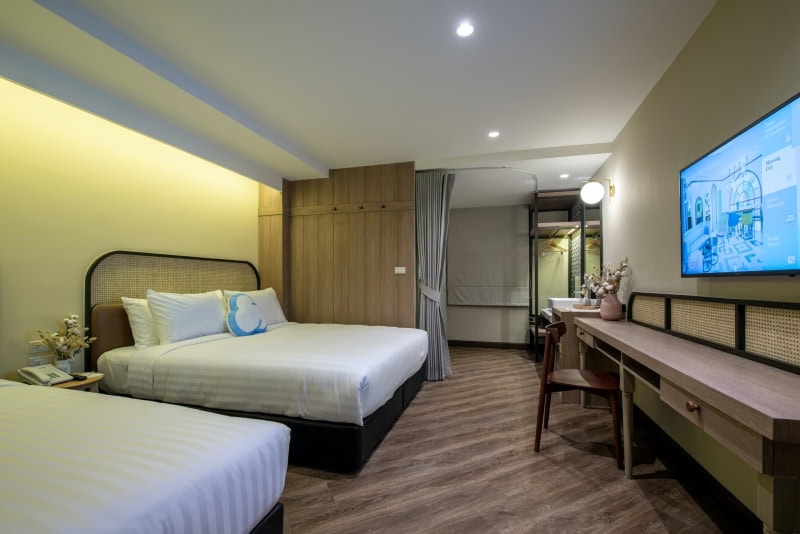 Modern hotel room with two double beds, wood flooring, a long desk with a chair, wall-mounted TV, soft lighting, and neutral decor. There is a mirror, flowers, and a curtain partially dividing the space.