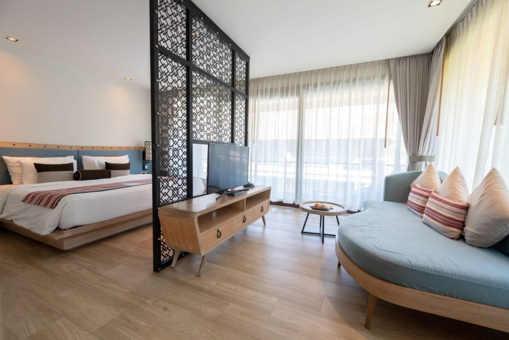 Modern hotel room with wooden floors, a large bed, a TV on a wooden console, and a blue cushioned sofa separated by a decorative black partition. Large windows with sheer curtains let in natural light.