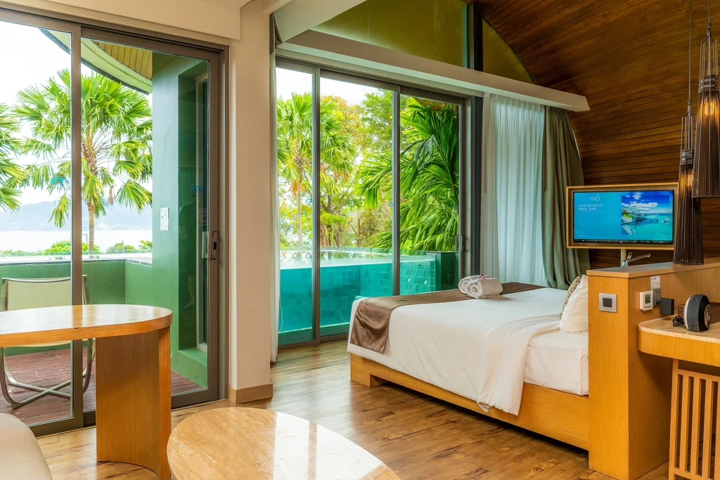 A modern hotel room with a bed, TV, round tables, wood flooring, and large glass doors opening to a patio with a small pool and lush tropical greenery outside.