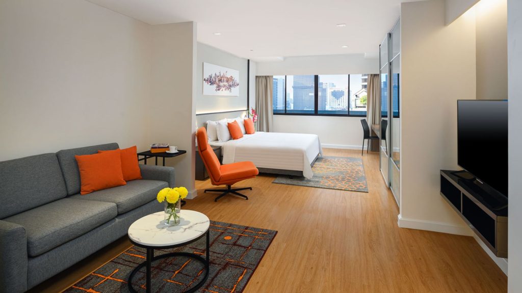 Modern hotel room with a bed, gray sofa with orange cushions, round coffee table with yellow flowers, wall-mounted TV, large window, and contemporary decor in neutral and orange tones.