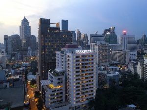 A cityscape at dusk featuring high-rise buildings, with the illuminated PARKROYAL hotel sign prominent in the foreground and various modern skyscrapers in the background under a blue evening sky.