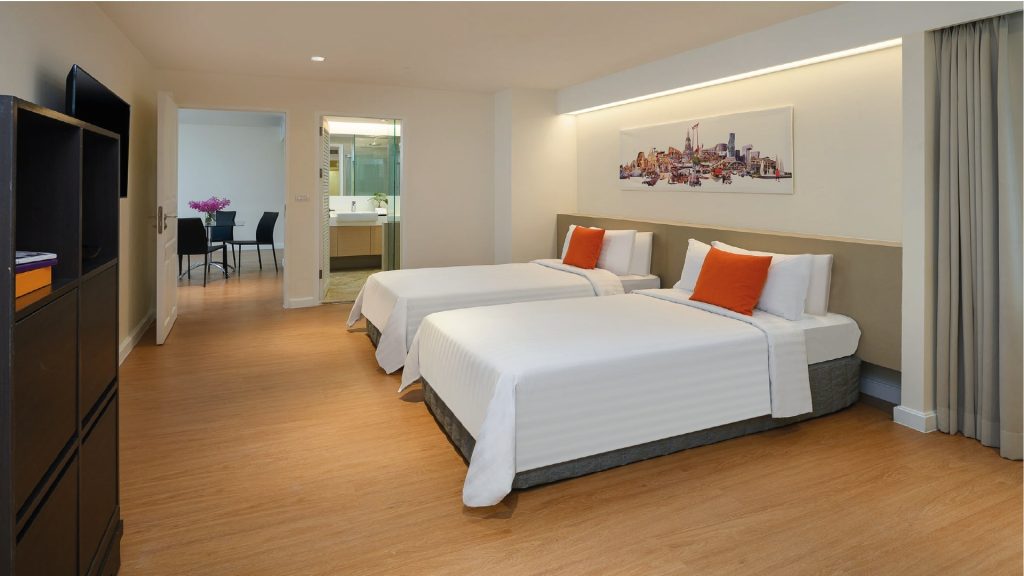 Modern hotel room with two neatly made twin beds featuring white linens and orange accent pillows, wooden flooring, a bathroom visible in the background, and a dining area with a table and chairs.